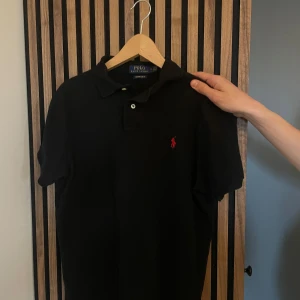 Ralph Lauren pike  - Säljer en klassisk svart pikétröja från Ralph Lauren med röd broderad logga på bröstet. Tröjan har korta ärmar och en slim fit passform. Perfekt för en stilren look. Modell 176, 57 kg 