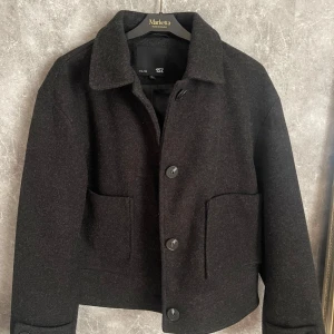 Mörkgrå ulljacka från 157 - Snygg mörkgrå jacka från 157 i ull med stora knappar framtill. Jackan är varm och skön med två stora fickor framtill. Perfekt för kyligare dagar. Kappan är helt felfri och har används 1 gång, den är som i nytt skick.