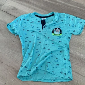 Turkos t-shirt från CMK Kids Club - Säljer en turkos t-shirt från CMK Kids Club med ett coolt tryck och knappar vid halsen. Perfekt för sommarens äventyr! 💙*Barnsize