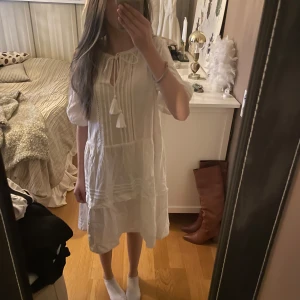 Vit klänning med knytband - Charmig vit klänning med knytband och volangdetaljer. Klänningen har trekvartsärmar och en luftig passform, perfekt för varma dagar.