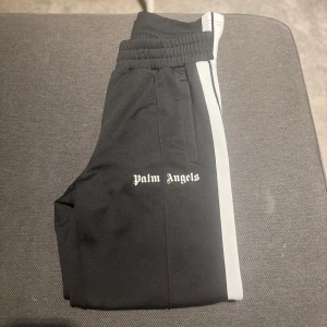 Svarta byxor från Palm Angels - Snygga svarta byxor från Palm Angels med vita sidoremsor. Byxorna har en elastisk midja och är perfekta för en avslappnad stil. Logotypen är tryckt på sidan för en extra touch av stil.