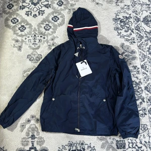 Mörkblå vindjacka från Moncler - Säljer en snygg mörkblå vindjacka från Moncler med huva och dragkedja. Jackan har långa ärmar och en stilren design med Moncler-logga på ärmen. Perfekt för blåsiga dagar!