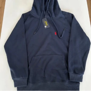 Ralph Lauren Hoodie - Säljer en helt ny och oanvänd, mörkblå hoodie från Polo Ralph Lauren med det klassiska röda logotypen på bröstet. Tröjan har en rymlig ficka framtill och justerbar huva med dragsko. Perfekt för en avslappnad stil.