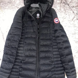 Svart dunjacka från Canada Goose - Säljer en svart dunjacka från Canada Goose med huva och dragkedja. Jackan har ett quiltat mönster och en logotyp på ärmen. Perfekt för kalla dagar.