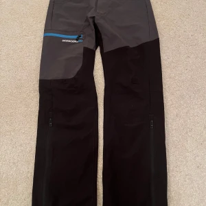 Svarta och grå byxor från Didriksons - Säljer ett par funktionella byxor från Didriksons i svart och grått. Byxorna har en praktisk dragkedja i blått och är perfekta för utomhusaktiviteter. Märket är broderat på sidan och de är designade för att skydda mot väder och vind.
