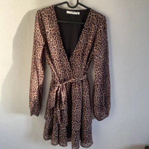 Leopardmönstrad klänning från NA-KD - Snygg leopardmönstrad klänning från NA-KD med v-ringning och långa ärmar. Klänningen har en omlott-design med knytband i midjan och volangdetaljer i nederkant. Perfekt för en trendig look! Ny och oanvänd stl 36. 