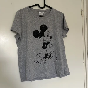 Grå t-shirt med Mickey Mouse från Disney - Grå t-shirt från Disney med en svart print av Mickey Mouse på framsidan. Klassisk rund halsringning och korta ärmar.