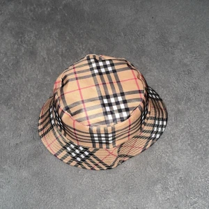 Beige rutig bucket hat från burberry  - Säljer en beige bucket hat med klassiskt rutigt mönster i svart, vitt och rött. Hatten har en mjuk form och passar perfekt för en trendig look.