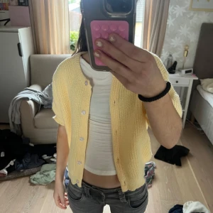 Gul stickad kofta med kort ärm - Säljer en ljusgul stickad kofta med korta ärmar och vita knappar framtill. Koftan har en avslappnad passform och är perfekt att slänga över en topp. Enkel och söt design som passar till många outfits.