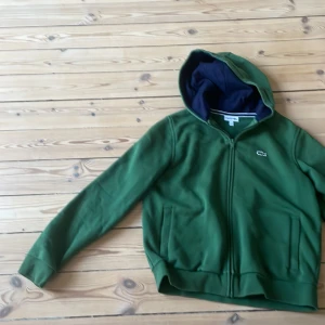 Grön hoodie från Lacoste Sport - Säljer en grön hoodie från Lacoste Sport med dragkedja och huva. Tröjan har långa ärmar, två fickor framtill och klassisk Lacoste-logga på bröstet. Perfekt för en sportig och avslappnad stil. Har haft på mig typ 5-10 gånger. Skriv bara vid funderingar!