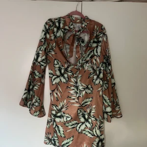 Blommig kortklänning med knytning - Säljer en kort klänning i satinliknande material med blommigt mönster i grönt och vitt på en beige botten. Klänningen har lång ärm, v-ringning och knytning framtill. Perfekt för sommarens tillfällen!