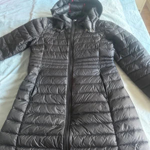 Svart lång dunjacka från JOTT - Säljer en svart lång dunjacka från JOTT med huva och dragkedja framtill. Jackan har ett quiltat mönster och en broderad logotyp på ärmen. Perfekt för kyliga dagar och har en smal passform. Köptes för 2700 kr, säljer för 1500 men pris kan diskuteras