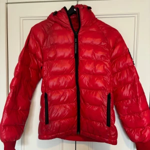 Röd dunjacka från Peak Performance - Säljer en röd dunjacka från Peak Performance med huva och svarta dragkedjor. Jackan har en quiltad design och två fickor framtill. Använd ett antal gånger men är hel och i helhet fin. Har ett svart/ grått märke på armen som syns på andra bilden. Går säkert bort vid tvätt som jag kan göra innan jag skickar iväg till köparen. 