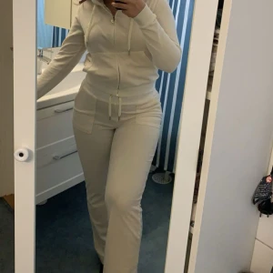 Vita mjukisbyxor från Juicy Couture - Säljer ett par klassiska vita mjukisbyxor från Juicy Couture med resår i midjan och dragsko. Byxorna har fickor med lock och knapp bak samt broderad logga framtill. Perfekta för en avslappnad och trendig look. Storlek S i byxan Storlek M i koftan