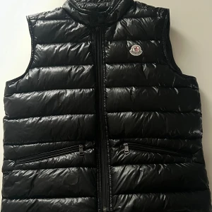 Moncler väst ”GUI” - Svart Moncler väst i storlek 2, På västen finns det 2 hål som vi nu har täckt över. På västen finns det också ett chip som man kan skanna för att direkt överföras till Moncler hemsidan.