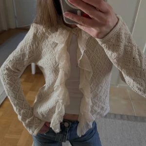 Beige spetskofta med volang - Supersöt beige kofta med spetsmönster och volang längs hela öppningen. Perfekt att slänga över en topp för en romantisk look.