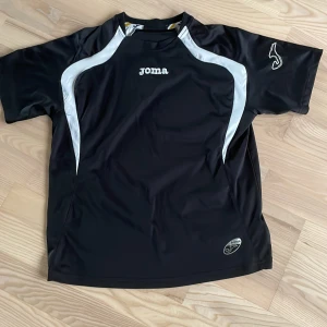 Svart sport t-shirt från Joma - Säljer en svart sport t-shirt från Joma med vita detaljer längs axlar och sidor. T-shirten har korta ärmar och rund hals. Perfekt för träning eller fotboll. Joma-logga på bröstet och ärmen.