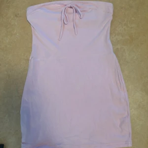 Ljusrosa tubklänning med knyt - Säljer en söt ljusrosa tubklänning med knyt framtill. Klänningen är axelbandslös och har en enkel, rak passform som passar perfekt till sommaren. Mjuk och bekväm, perfekt för varma dagar. Mer rosa i verkligheten, storlek M och bortklippta lappar