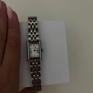 Snygg silverfärgad klocka från Mockberg - Elegant klocka från Mockberg i silverfärg med rektangulär urtavla och länkarmband i metall. Urtavlan är vit med romerska siffror och smala visare. Perfekt accessoar för en stilren look.