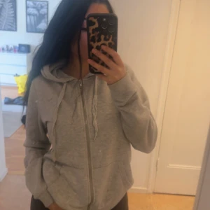 Grå hoodie med dragkedja - Grå hoodie med dragkedja och huva. Tröjan har långa ärmar och fickor framtill. 