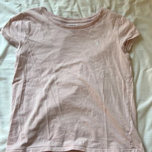Ljusrosa t-shirt från Polo Ralph Lauren - Ljusrosa t-shirt från Polo Ralph Lauren med klassisk rund halsringning och korta ärmar. T-shirten har en liten vit broderad logga på bröstet och är tillverkad i mjuk bomull. Perfekt för en enkel och stilren look.