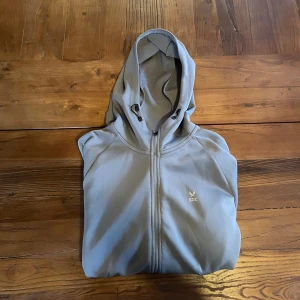 Ljusgrå hoodie med dragkedja från SOC - Ljusgrå hoodie med hel dragkedja och huva från SOC. Hoodien har två sidofickor med dragkedja och diskret logga på bröstet. Perfekt för träning eller chill. Mjuk och skön känsla med sportig look.