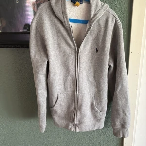 Grå hoodie med dragkedja från Polo Ralph Lauren - Säljer en klassisk grå hoodie från Polo Ralph Lauren i barn storlek XL. Tröjan har dragkedja framtill, två fickor och huva. Liten broderad logga på bröstet. Perfekt för en avslappnad stil.