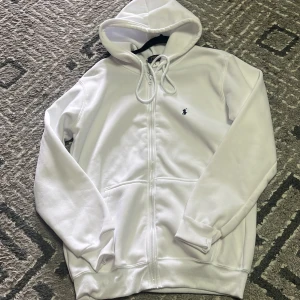 Vit hoodie med dragkedja från Ralph Lauren - Säljer en vit hoodie från Ralph Lauren med dragkedja framtill och huva med snörning. Tröjan har en liten broderad logga på bröstet och ribbade muddar vid ärmslut och nederkant.