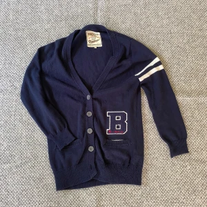 Mörkblå kofta med broderad B från Bondelid - Säljer en mörkblå kofta från Bondelid med broderad B på bröstet och två vita ränder på ena ärmen. Koftan har knappar framtill, ribbade muddar och två fickor. Perfekt för en sportig och avslappnad stil.