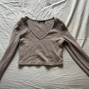 Beige croppad långärmad topp med v-ringning - Trendig beige långärmad topp med croppad passform och v-ringad hals. Tillverkad i ett ribbat material med diskret struktur. Perfekt för en stilren och avslappnad look.