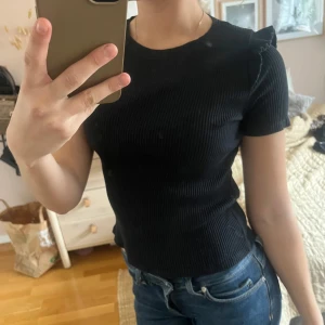 Svart ribbad topp från Zara - Säljer en svart ribbad topp från Zara med korta ärmar och rund halsringning. Toppen har fina volangdetaljer på axlarna och är figurnära i modellen. Perfekt att styla med jeans eller kjol.