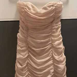 Ljusbeige draperad klänning från H&M - Supersnygg ljusbeige kortklänning från H&M med draperad design och axelbandslös passform. Perfekt för dig som vill sticka ut med en elegant och trendig look. Klänningen har en figurnära siluett och mjukt, syntetiskt material.
