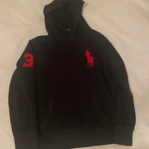  Polo Ralph Lauren hoodie - Säljer en svart hoodie från Polo Ralph Lauren med röd broderad logga på bröstet och en röd siffra 3 på ärmen. Klassisk känguruficka framtill och mjukt material. Perfekt för en avslappnad och stilren look.