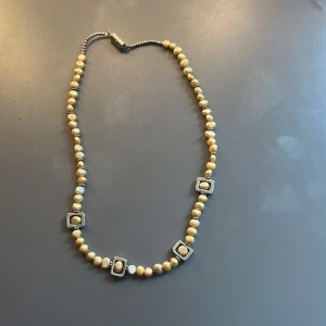 Guldfärgat pärlhalsband med fyrkantiga detaljer - Handgjort halsband med guldfärgade pärlor och fyra fyrkantiga detaljer i silverfärg runt om. Pärlorna är runda och skimrande, och halsbandet har ett metalliskt magnetlås. 