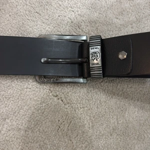 Svart bälte från Diesel med silverdetaljer - Säljer ett svart skärp från Diesel med silverfärgat spänne och märkets logga ingraverad. Skärpet har en stilren design och en metallnit som detalj vid fästet. Perfekt accessoar för att lyfta din outfit.