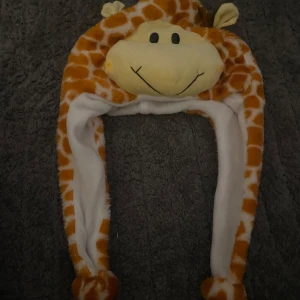 Giraffmössa med öron och ansikte - Rolig och mjuk mössa i form av en giraff med öron och broderat ansikte. Mössan har ett orange och vitt giraffmönster och långa hängande sidor. Perfekt för att sticka ut eller till temafest.