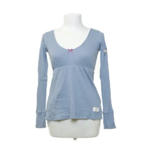 Ljusblå långärmad topp från Odd Molly - Supersöt ljusblå långärmad topp från Odd Molly med v-ringning och liten rosa rosett framtill. Tröjan har ribbad struktur och spetsdetalj vid halsringningen. Perfekt för dig som gillar en feminin och avslappnad stil.