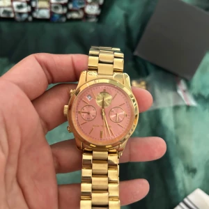 Guldig klocka med rosa urtavla från Michael Kors - Säljer en snygg klocka från Michael Kors i guld med rosa urtavla. Klockan har kronograf-funktion, datumvisning och kommer med extra länkar. Perfekt accessoar för att lyfta din stil.