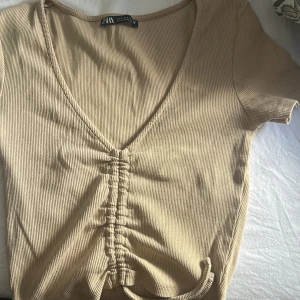 Beige ribbad topp -  beige ribbad topp från Zara med v-ringning och knyt framtill. Toppen har korta ärmar och en snygg rynkdetalj längs mitten! Super fin