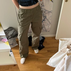 Olivgröna cargopants med raka ben - Olivgröna cargopants med raka/vida ben och flera fickor på sidorna. Byxorna har resår i midjan och dragkedja framtill. I bra skick. Strl 36 (jag har normalt 34). 