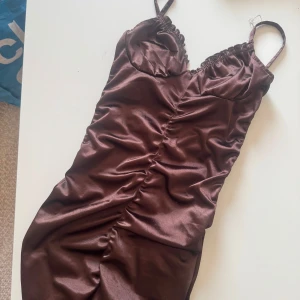 Brun satin klänning från Divided - Snygg brun kortklänning i satin från Divided med smala axelband och rynkade detaljer framtill. Klänningen har en figurnära passform och en lätt glansig yta som ger en lyxig känsla. 