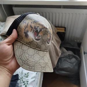 Beige keps med tigertryck från Gucci - Säljer en beige keps från Gucci med klassiskt GG-mönster och ett stort tigertryck framtill. Kepsen har svart mesh baktill och justerbar rem med röd-gröna detaljer. Perfekt för dig som vill sticka ut med en lyxig accessoar.