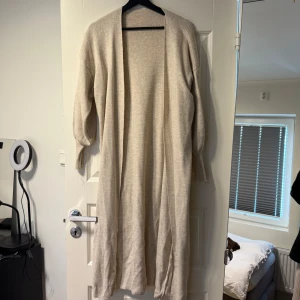 Beige lång kofta i mjukt material - Lång, öppen beige kofta med avslappnad passform och långa ärmar. Perfekt att slänga över en outfit för extra värme. Mjuk och skön känsla, passar till många stilar.