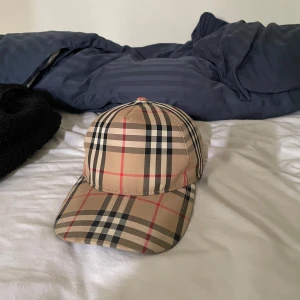 Beige rutig keps från Burberry - Snygg keps från Burberry i klassiskt beige rutigt mönster med svarta, vita och röda linjer. Justerbar baktill med svart spänne och broderad Burberry-logga. Perfekt accessoar för en trendig look. kan även vara öppen för byten 