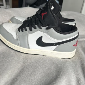 Nike Air Jordan 1 Low i grått och vitt - Säljer ett par Nike Air Jordan 1 Low i grått och vitt med svarta detaljer och röd logga på hälen. Skorna har svart snörning, svart swoosh och klassisk rund tå. Perfekta för dig som gillar streetwear och sneakers.