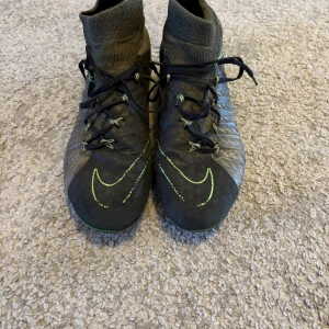 Nike hypervenom🐊🐊 - Säljer ett par svarta fotbollsskor från Nike med gröna detaljer och högt skaft. Skorna har snörning och är tillverkade i syntetmaterial med textil vid ankeln för extra stöd. Perfekta för dig som vill ha bra grepp och stil på planen.