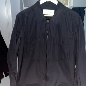 Svart vindjacka från A Day's March - Säljer en stilren svart vindjacka från A Day's March i storlek S. Jackan har dragkedja framtill, två bröstfickor med lock och klassisk krage. Perfekt för dig som gillar minimalistisk design och lätta material.