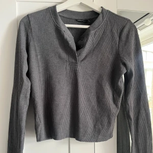 Mörkgrå ribbad långärmad topp - Säljer en mörkgrå ribbad långärmad topp med knappar vid halsringningen. Modellen är croppad och har en enkel, stilren design som passar till många olika outfits. Aldrig använd. Skriv för fler bilder💞