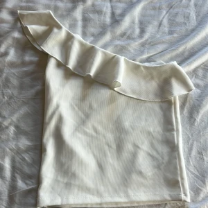 Vit oneshoulder topp med volang från Stradivarius - Säljer en vit ribbad oneshoulder topp från Stradivarius med bred volangdetalj längs axeln. Snygg och trendig modell som passar perfekt till sommaren. Materialet är mjukt och stretchigt. Har aldrig använts.