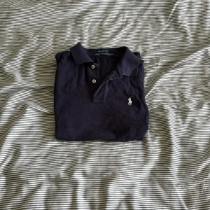 Svart pikétröja från Ralph Lauren - Säljer en klassisk svart pikétröja från Ralph Lauren med korta ärmar och broderad logga på bröstet. Perfekt för en stilren och avslappnad look. Passar bra till både jeans och chinos.